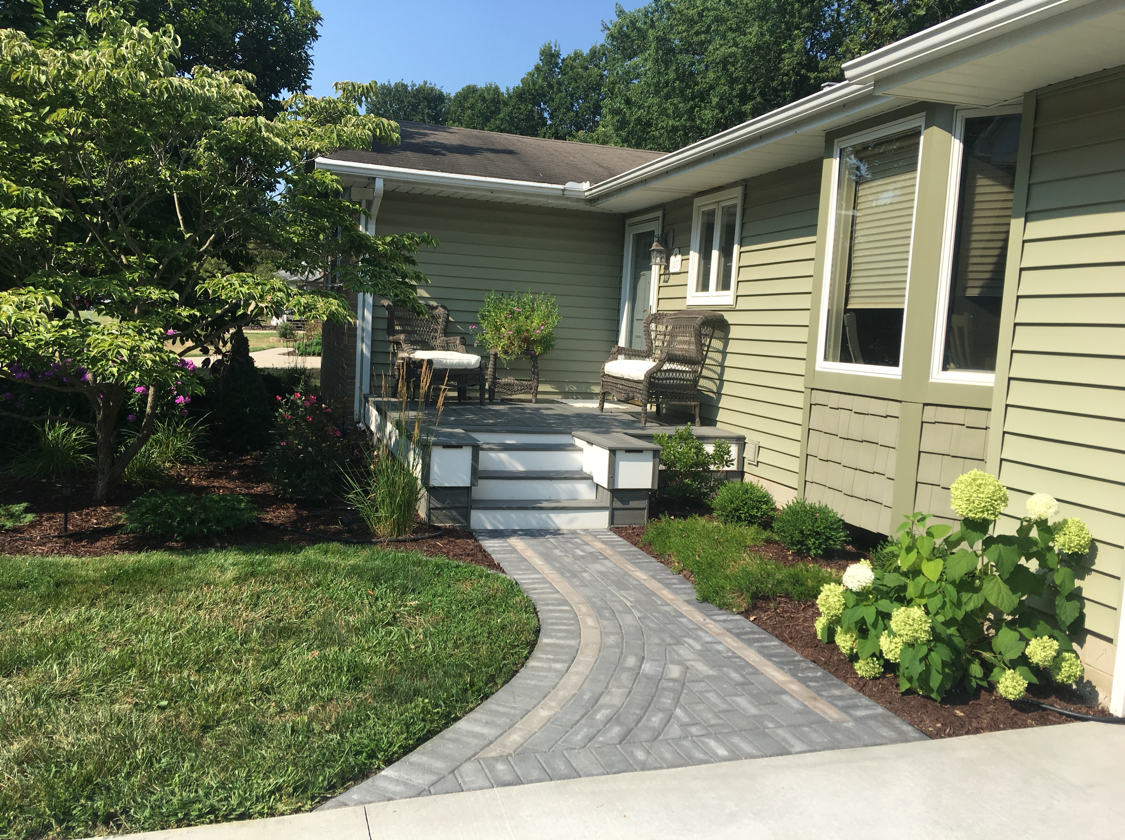 Paver pathway leads to a house with sage green siding, small porch with chairs, and landscaping.