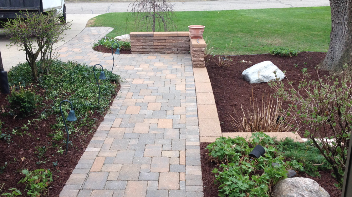Brick walkway leading to a garden with green plants and mulch beds.