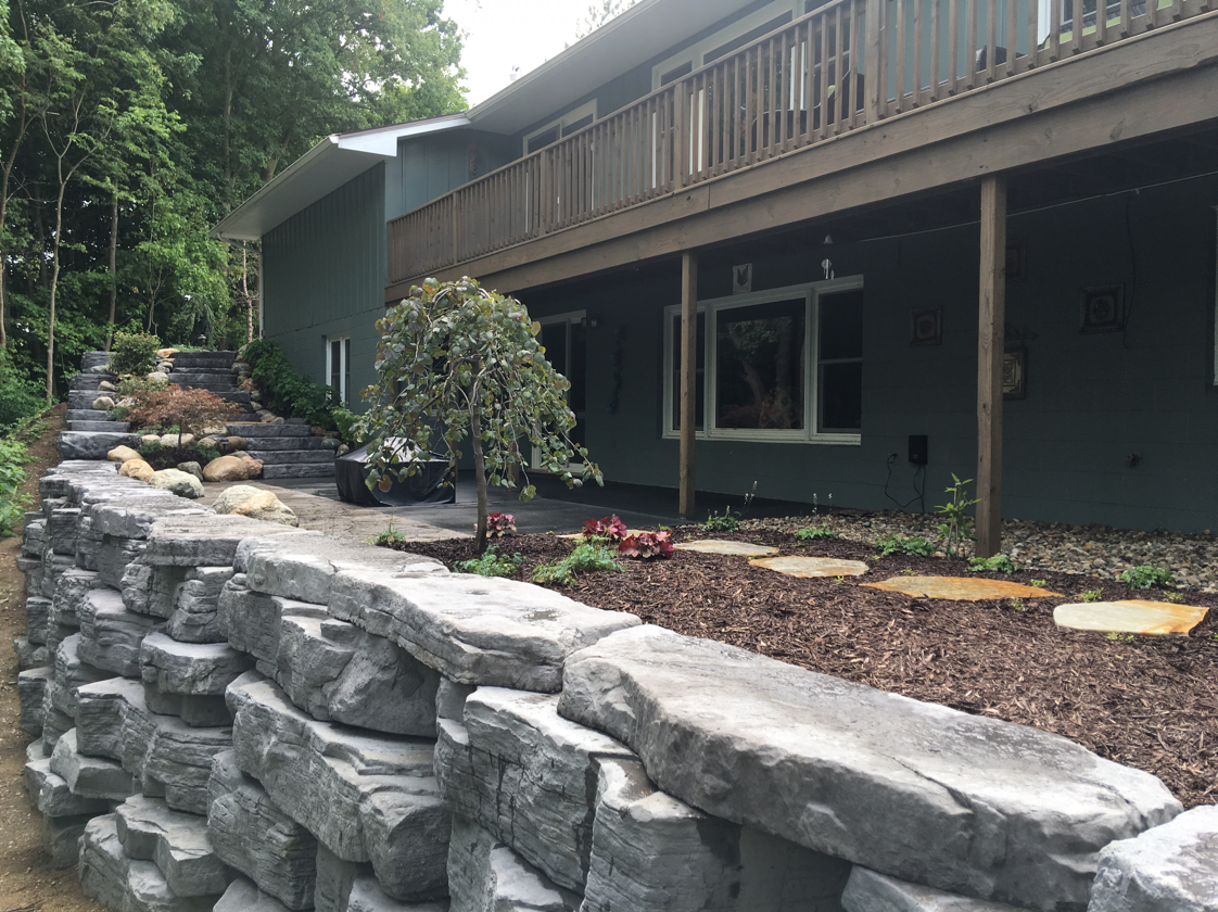 Stone retaining wall supports landscaped yard alongside a green house with a wooden deck.