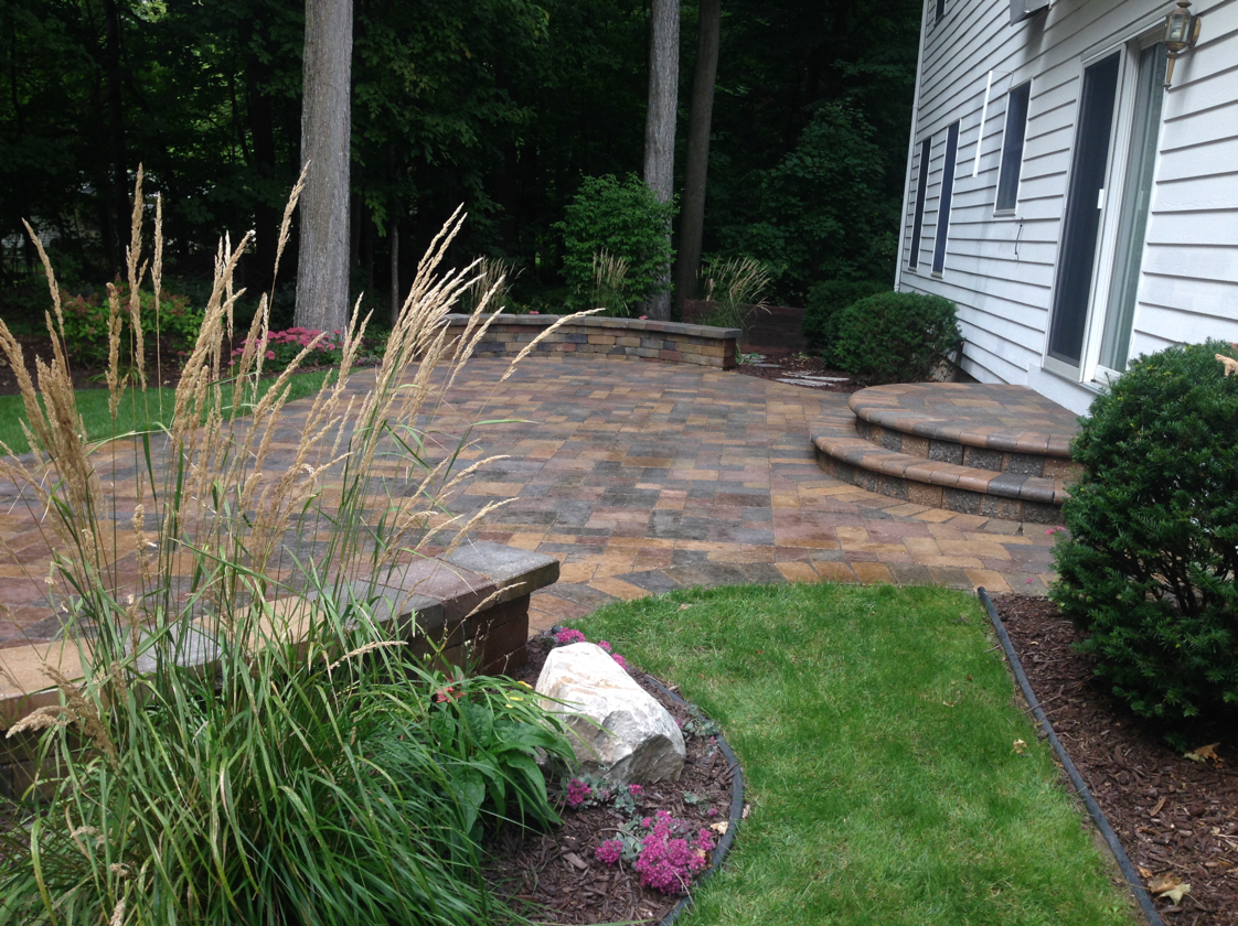 Brick patio with steps, bordered by green grass and plants, next to a white house.