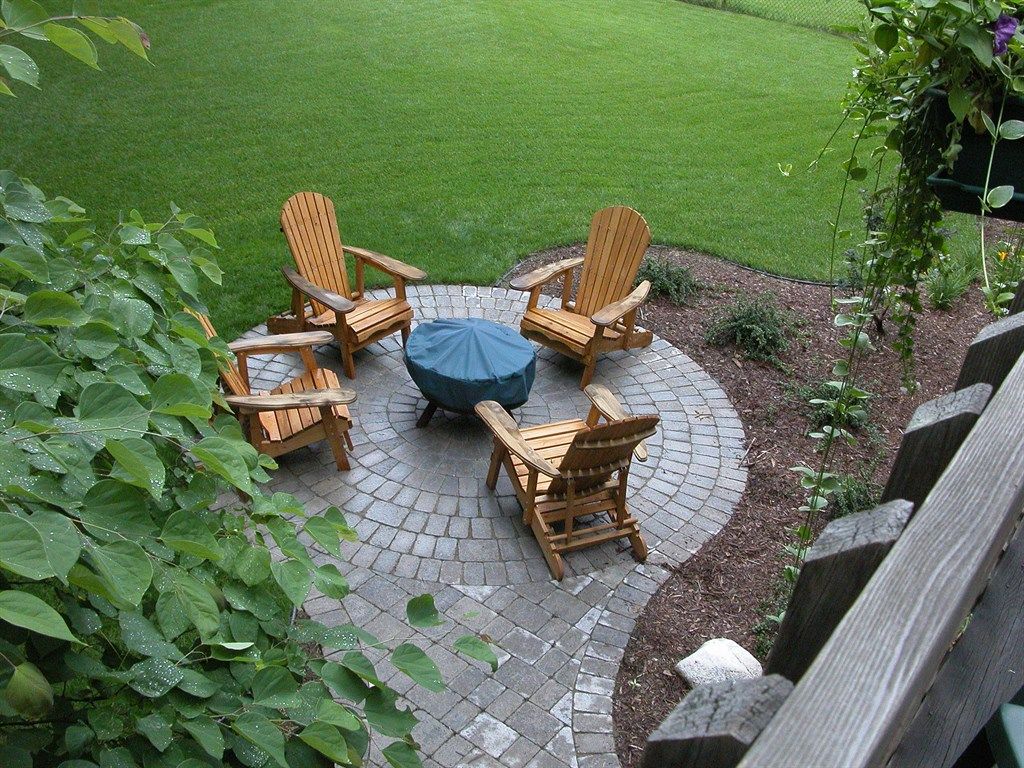 Four wooden chairs surround a fire pit on a brick patio, green grass in the background.