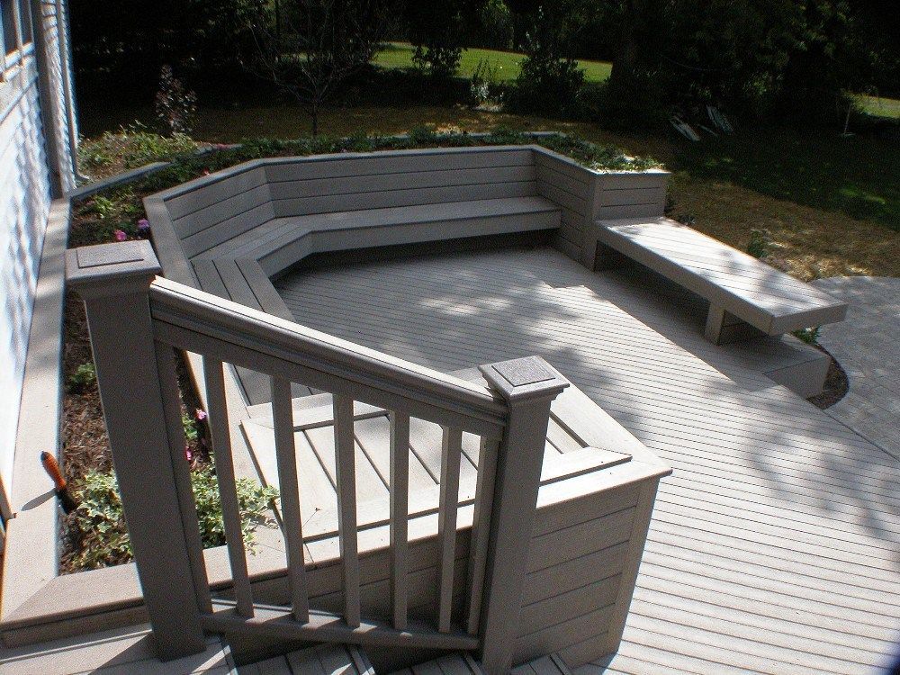 Gray deck with built-in seating area. Low railing in foreground, grass, and trees in the background.