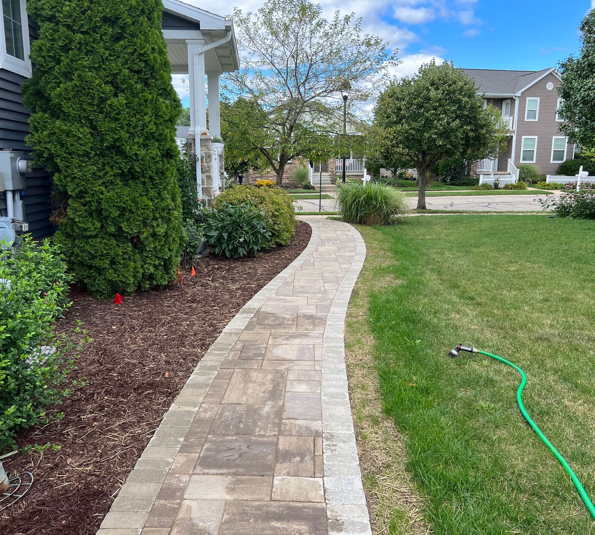 A brick pathway curves through a green yard and garden bed, leading to a house with a porch.