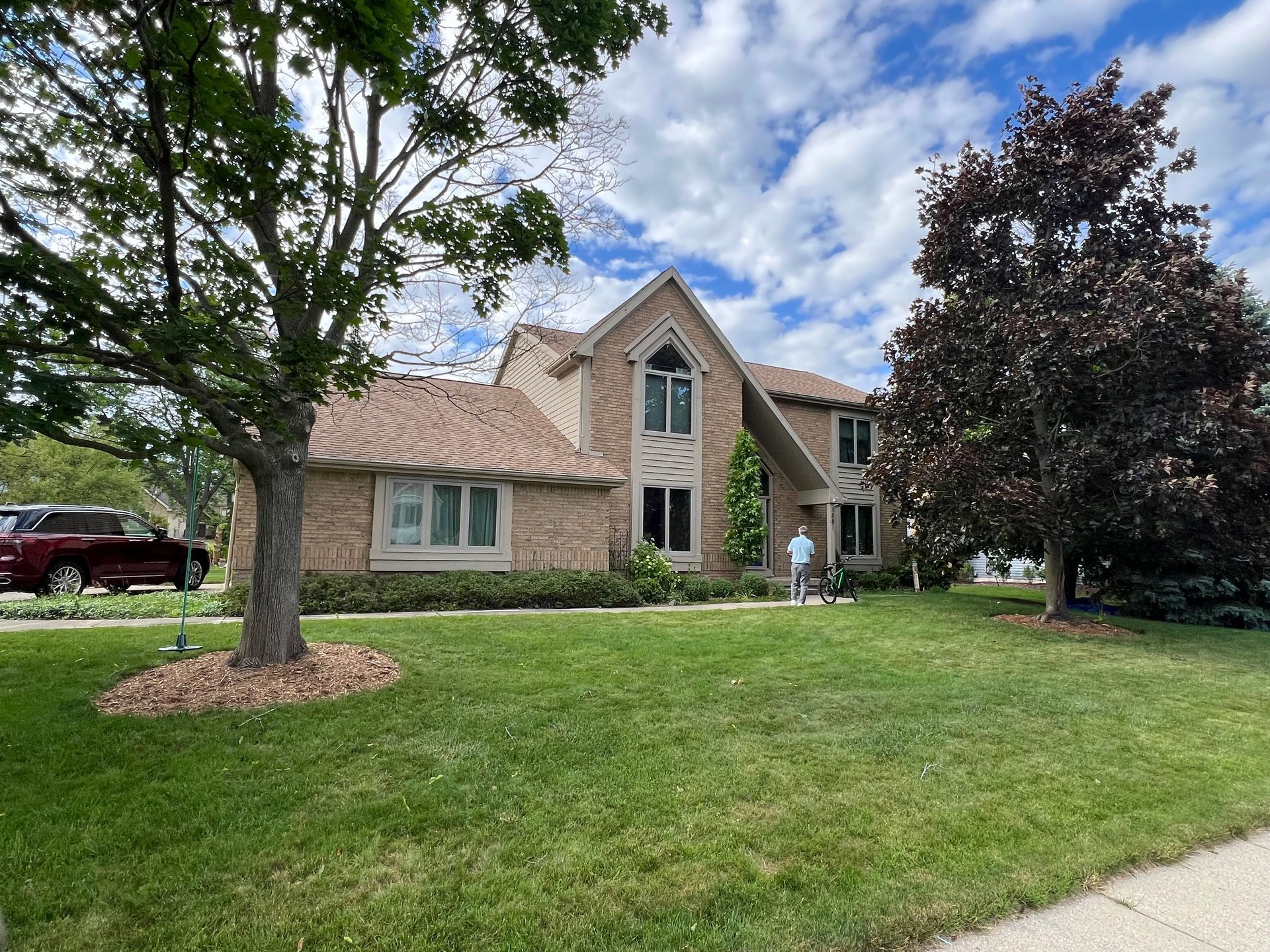 Two-story brick house with green lawn, trees, and cloudy sky; person standing in front.