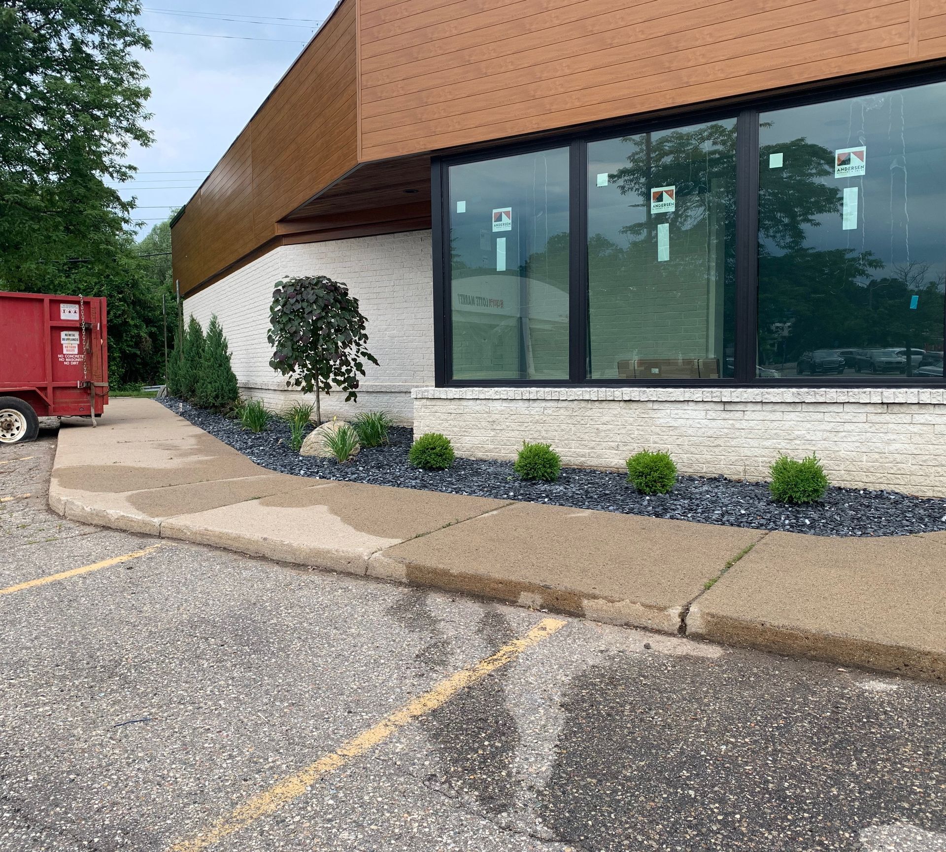 Exterior view of a building with a concrete sidewalk, landscaping, and windows. A red dumpster is on the left.