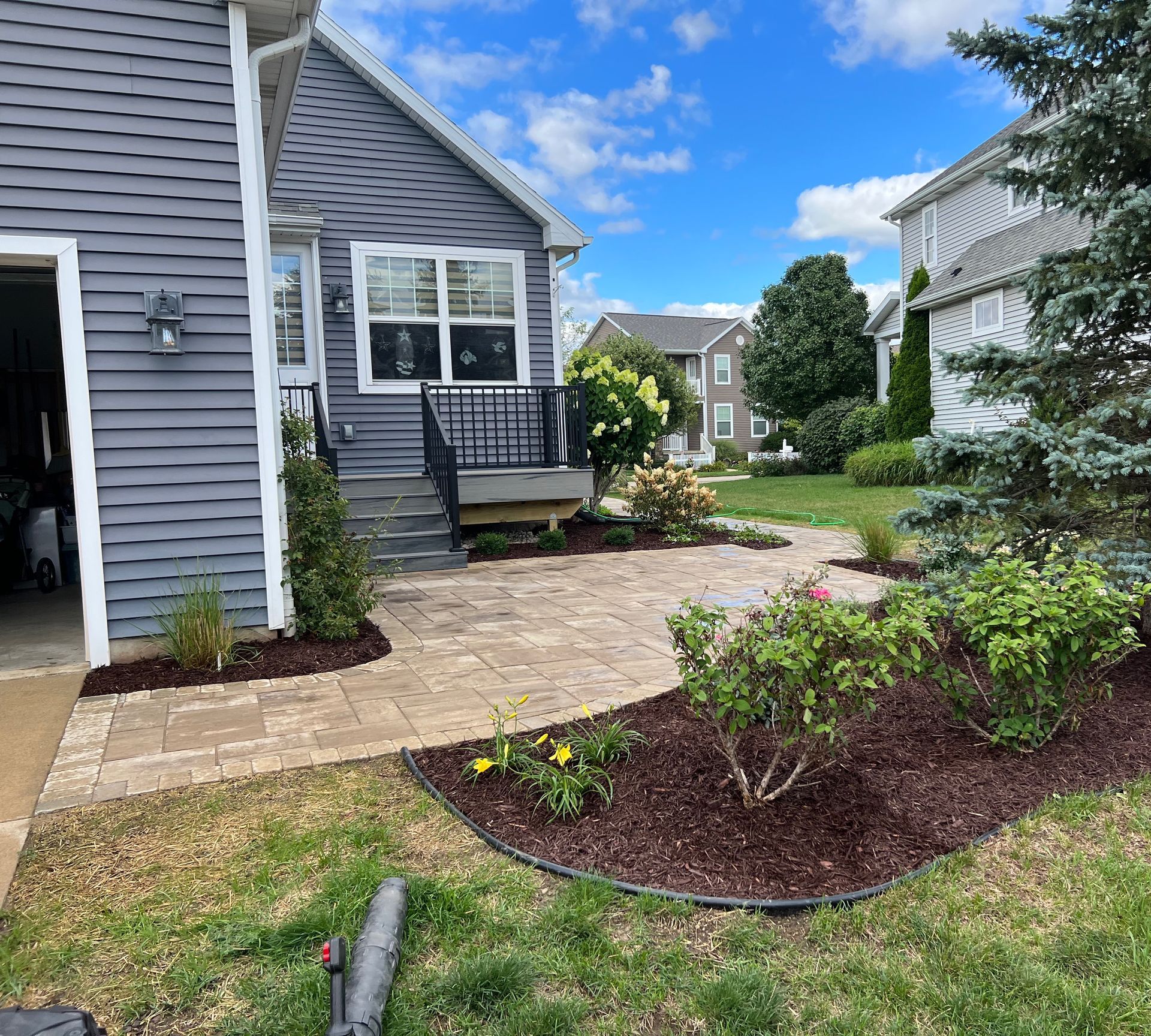 Backyard patio with landscaping, house, and green lawn. Blue sky, neutral-toned pavers, brown mulch, and various plants.