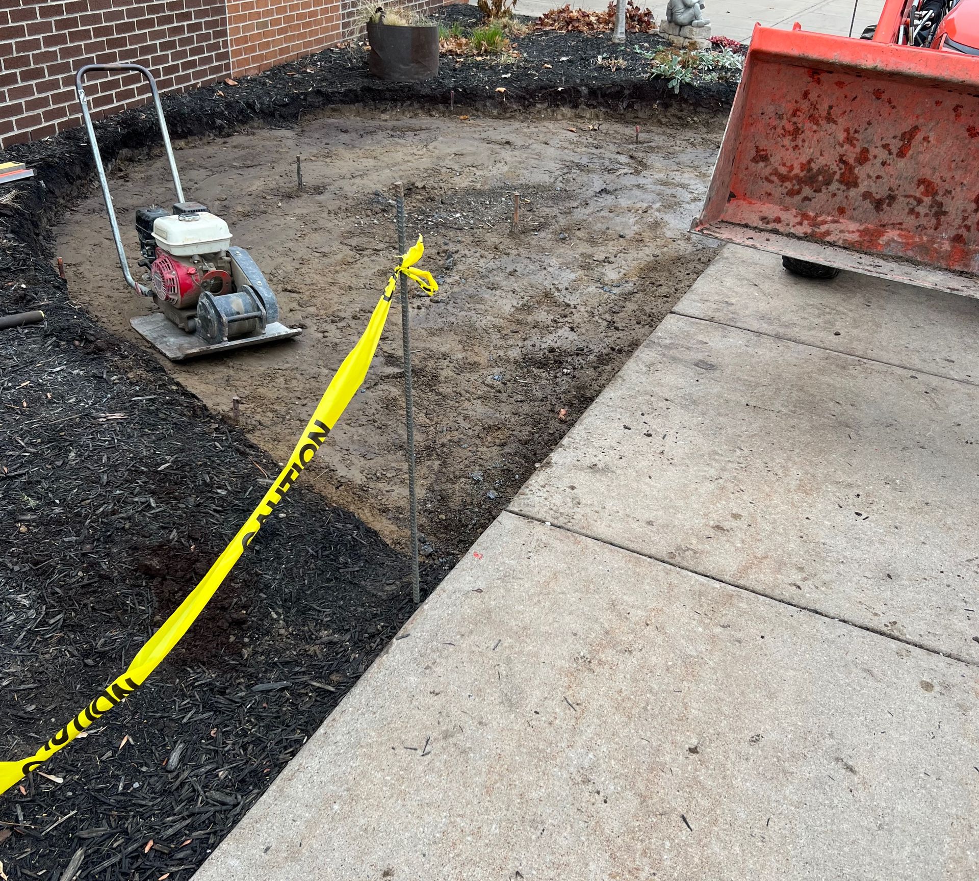 A plate compactor sits on a patch of cleared dirt next to a concrete sidewalk, with caution tape and a skid steer bucket.