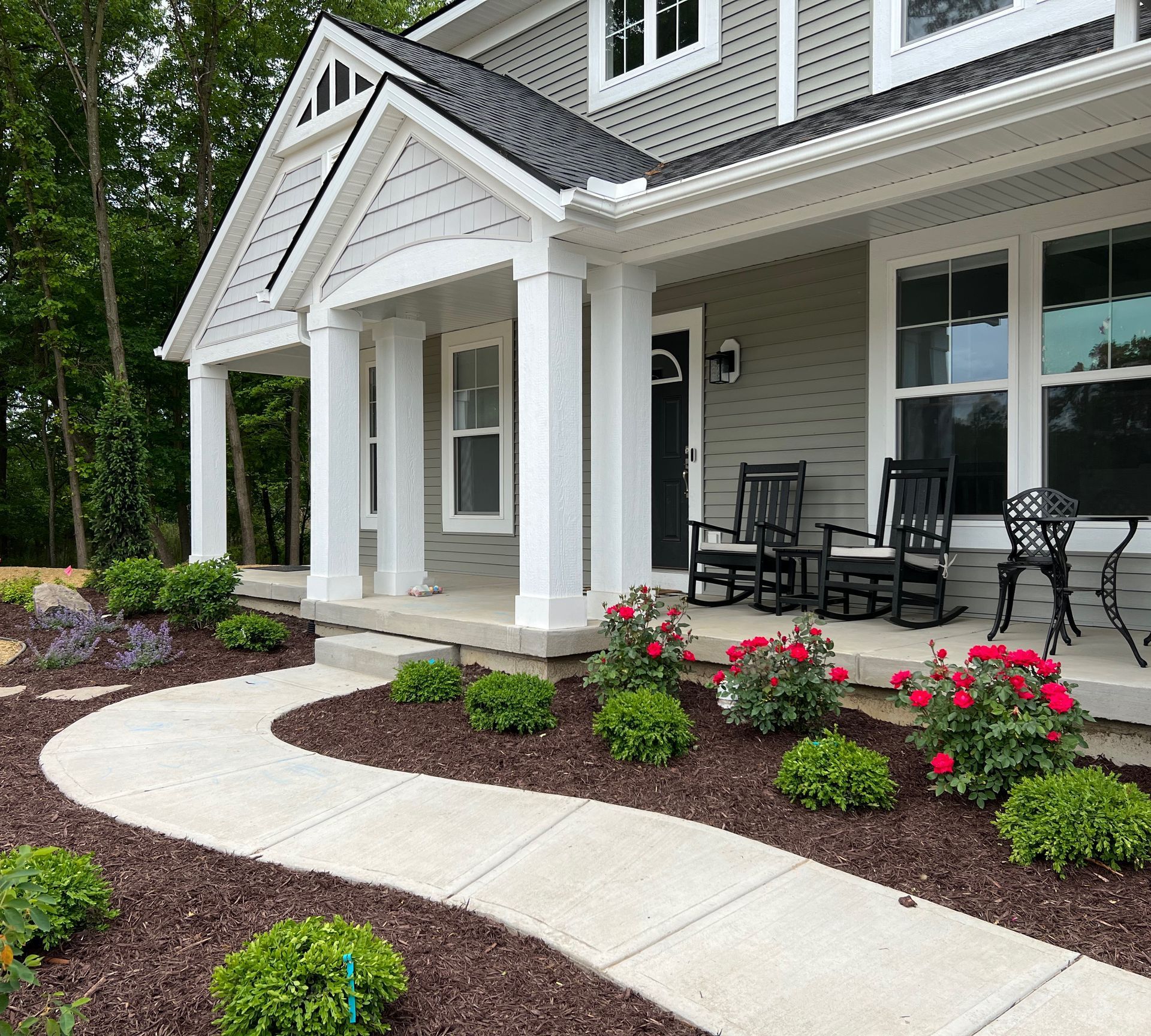 Gray house with a front porch. A winding concrete walkway is lined with bushes and flowers.