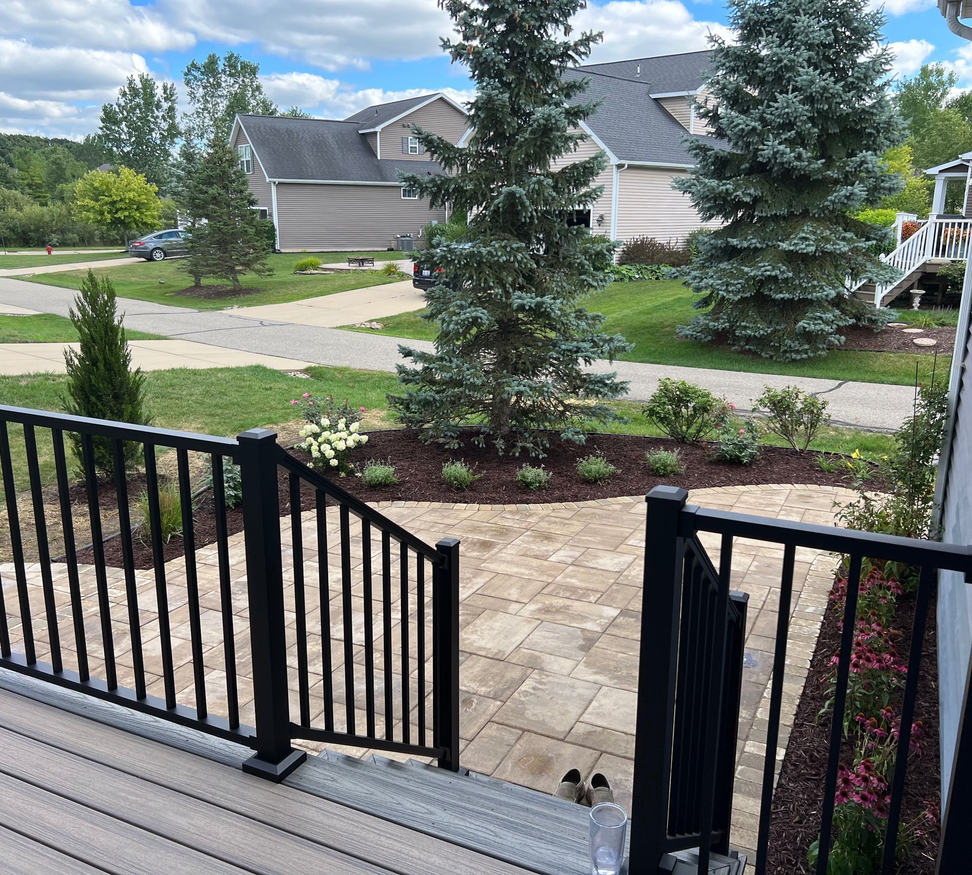 View from a porch with black railing overlooking a landscaped yard, stone patio, and neighborhood street.