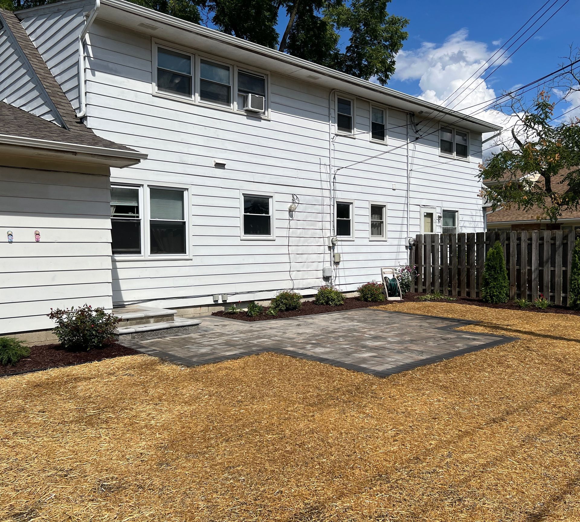 Backyard with a two-story white house, a paved patio, and wood chip ground cover under a blue sky.