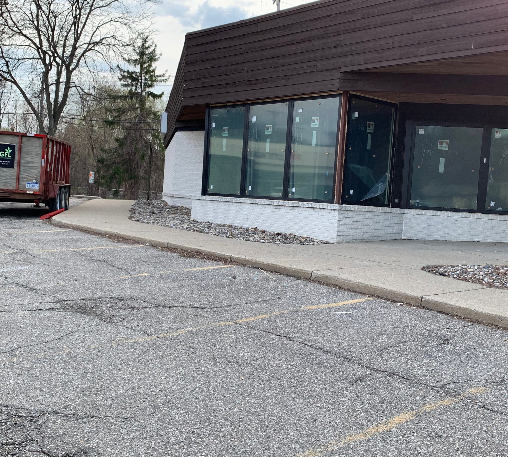 Exterior of building with large windows, brown roof, and cracked asphalt parking lot. A dumpster sits nearby.