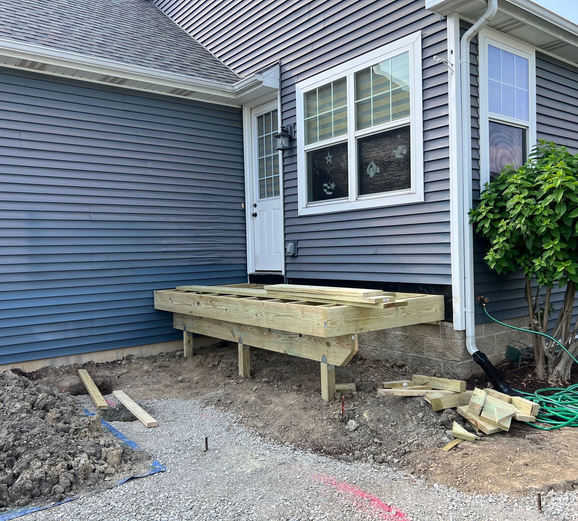 Wooden deck under construction next to a house with blue siding. Gravel path and some tools visible.