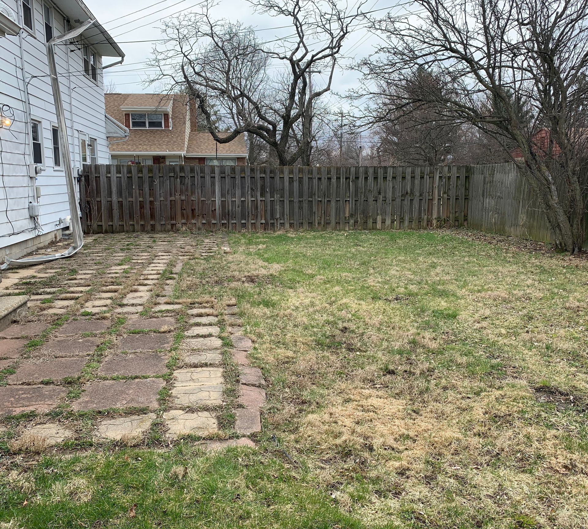 Backyard with brick patio, patchy grass, wooden fence, and bare trees.