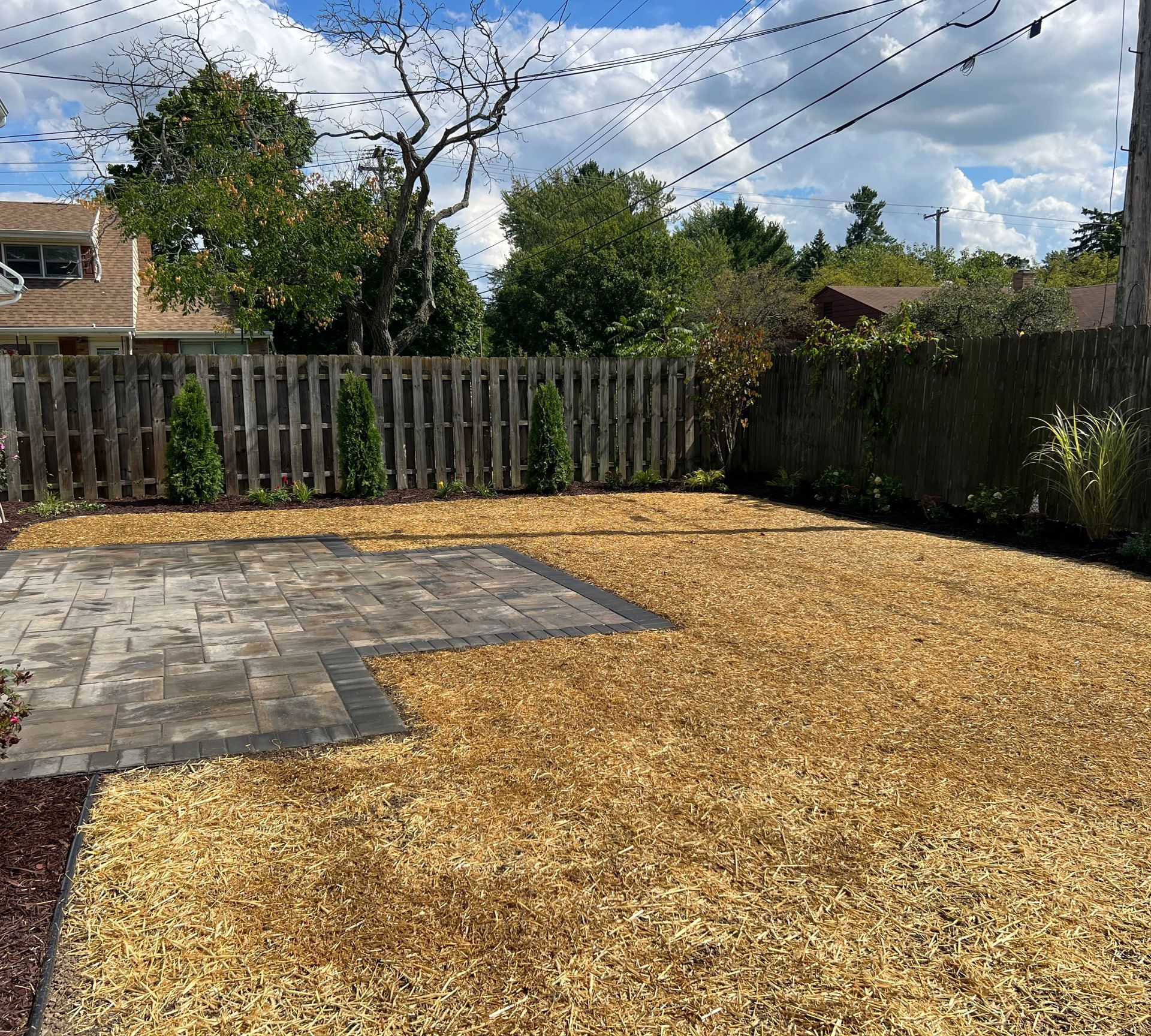 Backyard with wood chip ground cover, brick patio, and wooden fence under a cloudy sky.