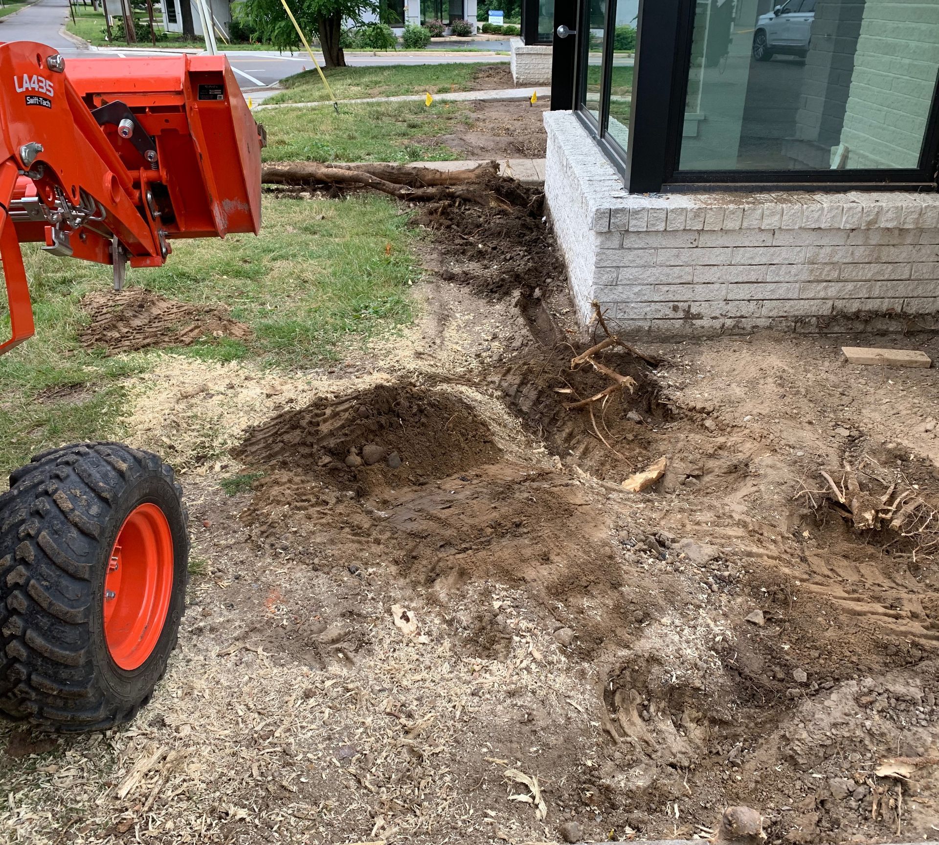 Orange tractor digging near a building with a large window.  Dirt and wood chips cover the ground.