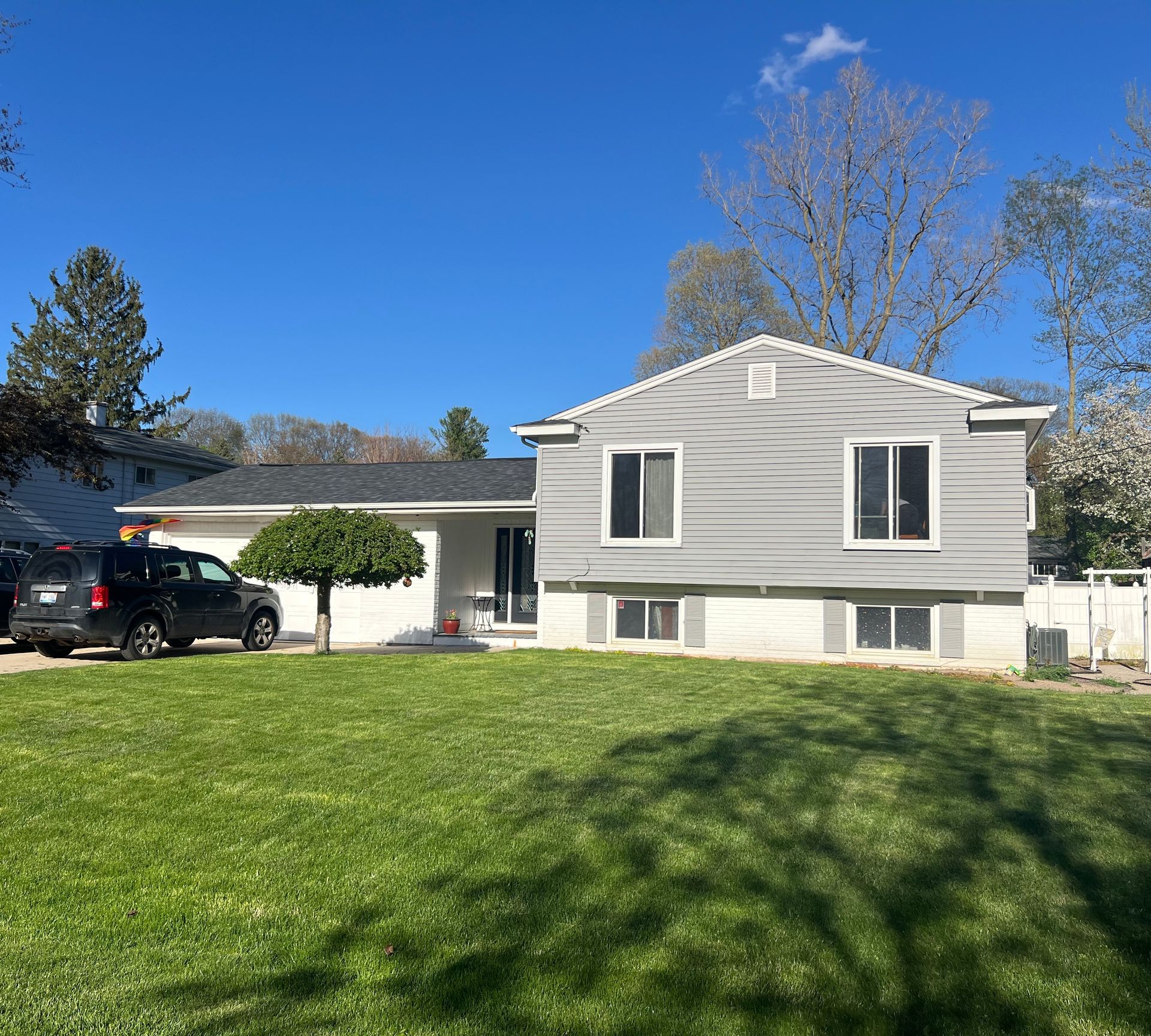 Gray house with black car in the driveway on a sunny day. Green lawn and blue sky.