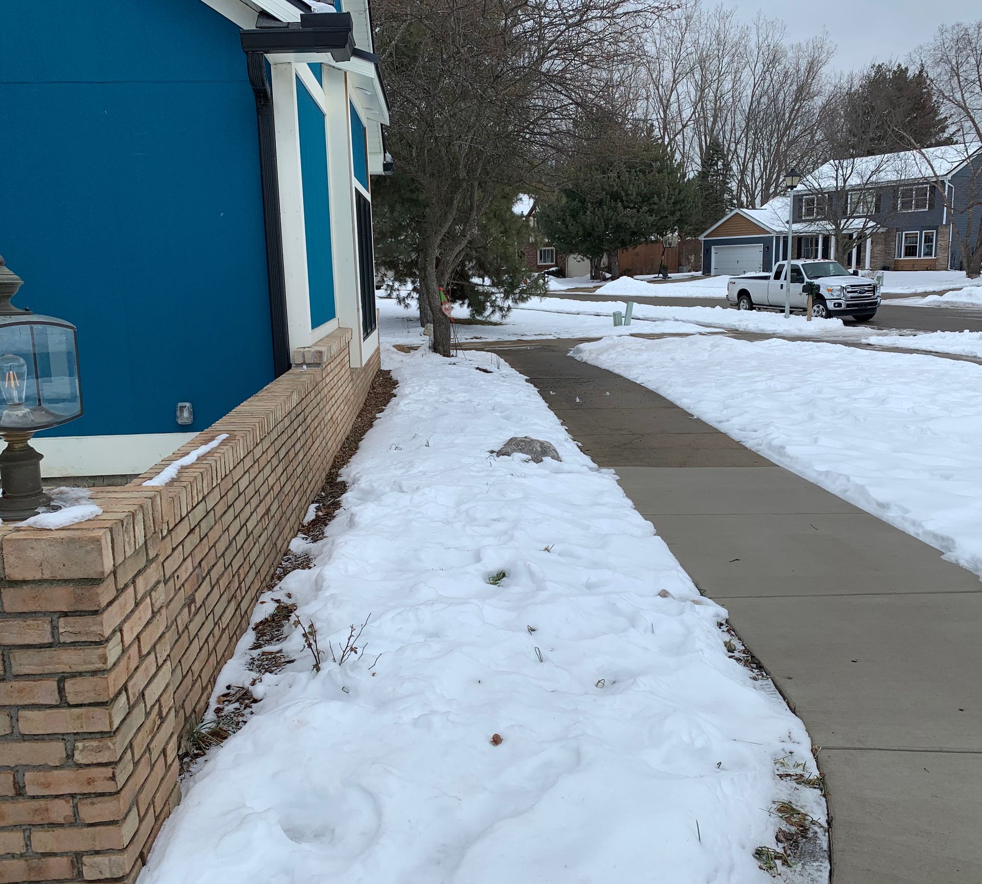 Snowy residential street; a blue house with brick wall borders a snow-covered sidewalk.