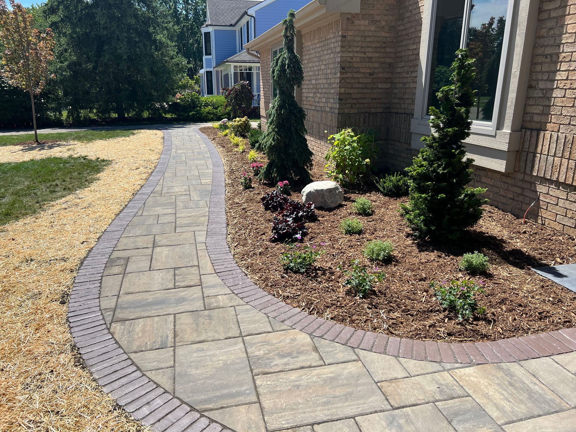 Brick pathway curves past a flower bed with mulch, stone and greenery, next to a home with brick siding.
