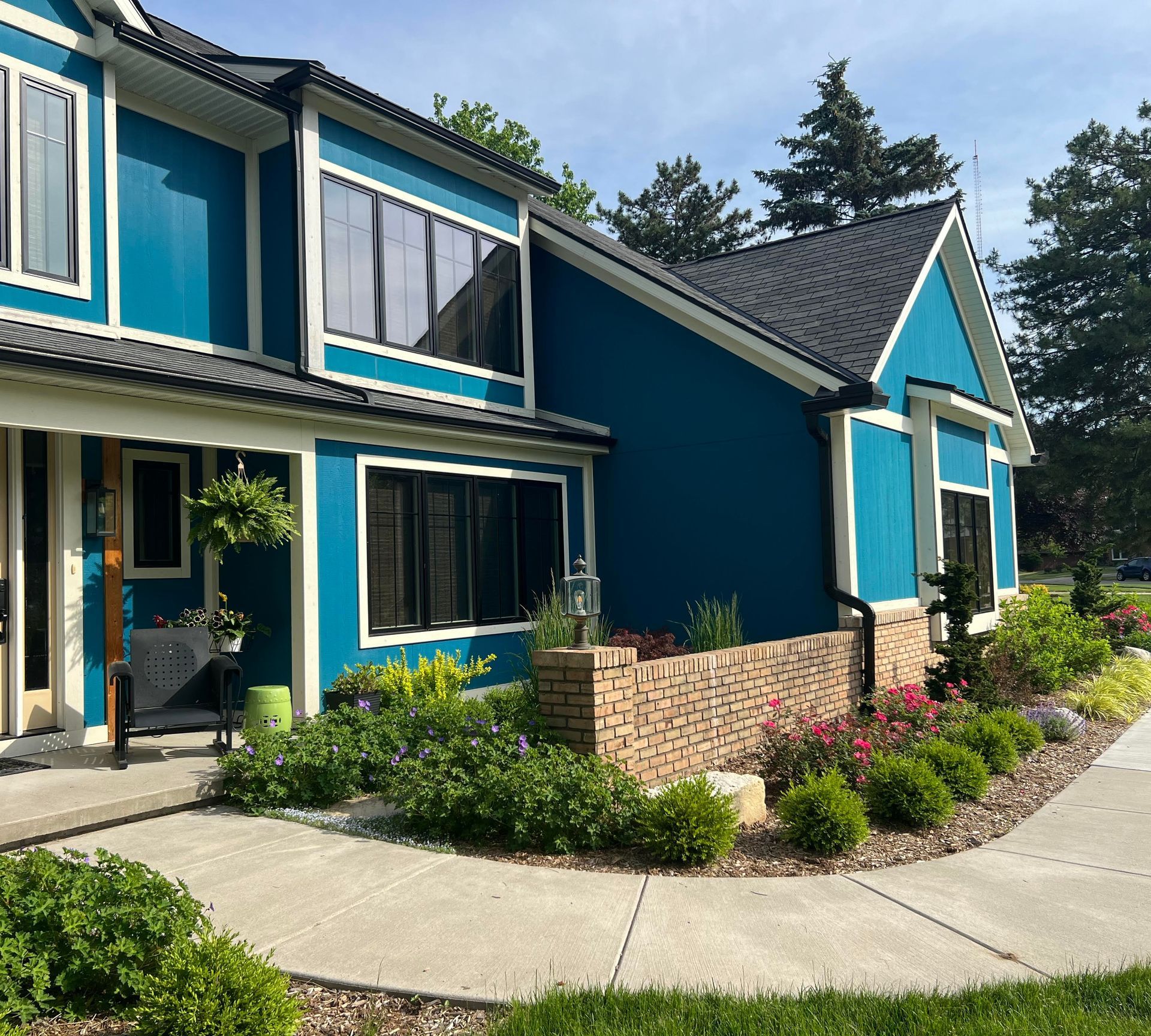 Blue house with black trim and a black roof, next to a curved brick wall and sidewalk. Lush landscaping.