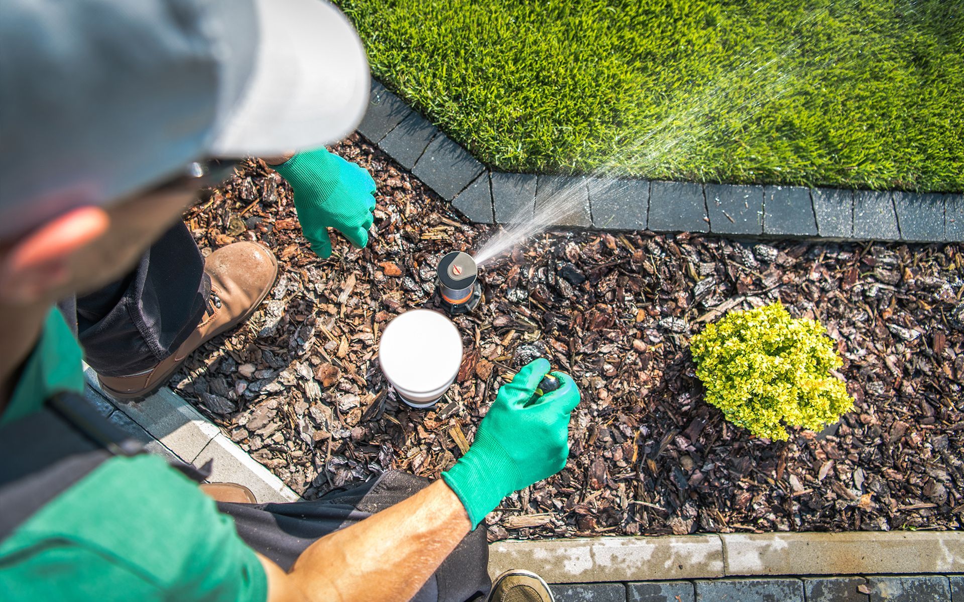 Person adjusting a sprinkler head, wearing gloves, next to mulch and a green lawn.