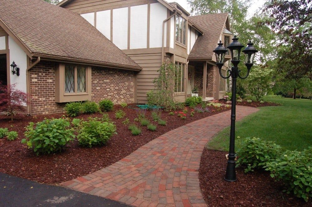 Brick pathway leads to a house with a brown roof and landscaped yard with dark mulch and green bushes.