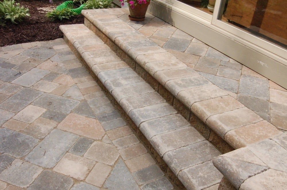 Stone steps leading up to a building, with a patterned brick patio in varying shades of tan and gray.