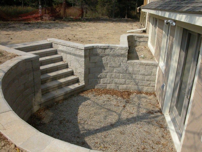 Exterior basement access with concrete steps and retaining walls. Gravel ground, building on the right.