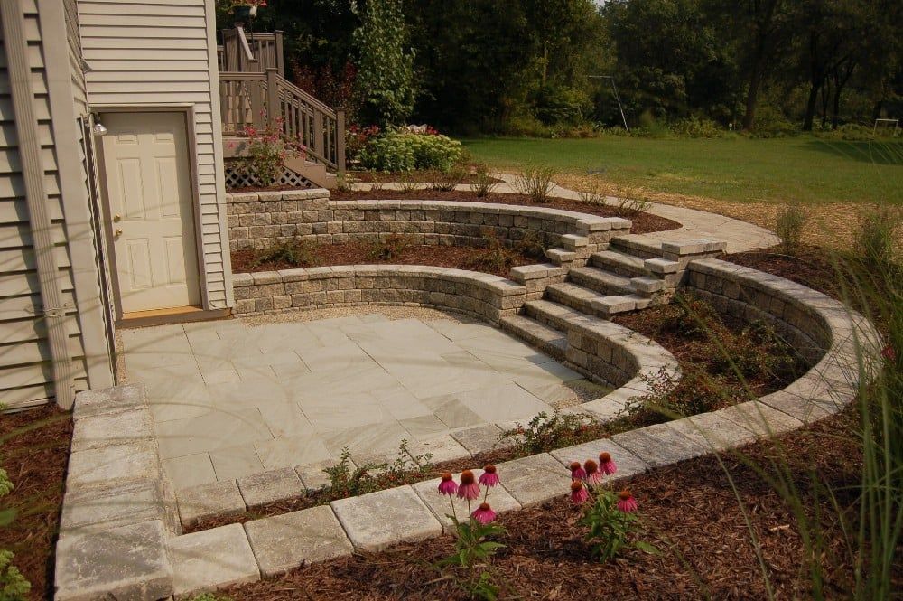 Stone patio with steps and retaining walls next to a house, leading to a grassy area.