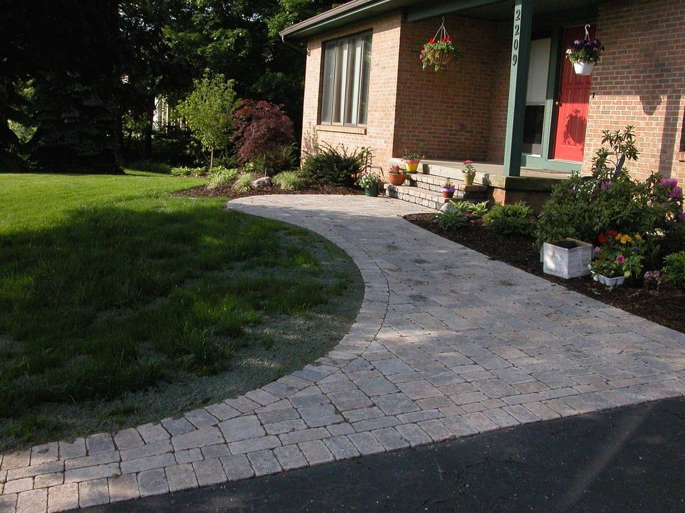Brick walkway curving from dark asphalt driveway to a brick house with a red door and flower pots.