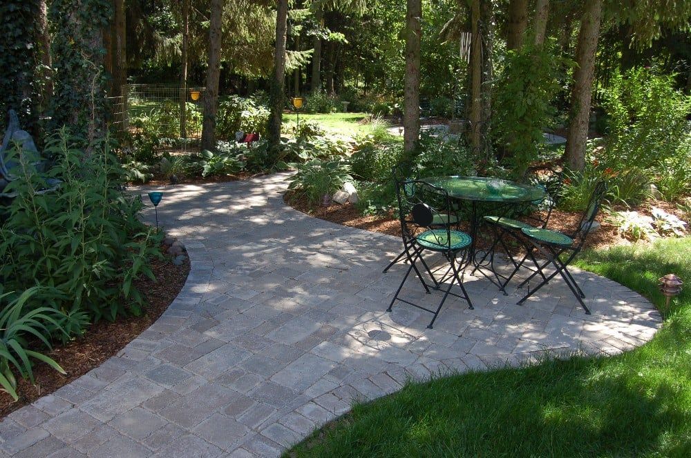 Brick pathway winds through a lush garden to a table and chairs, surrounded by trees and green lawn.