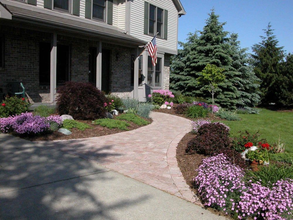 Brick pathway leads to a house with a flag; pink and purple flowers border the path.