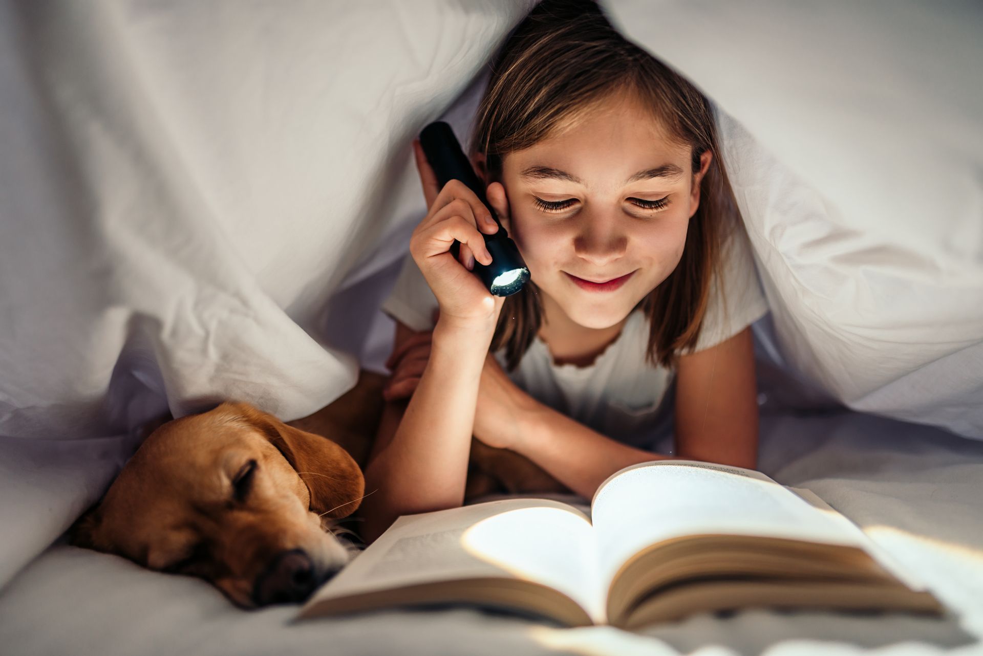 A little girl is reading a book to her dog under a blanket.