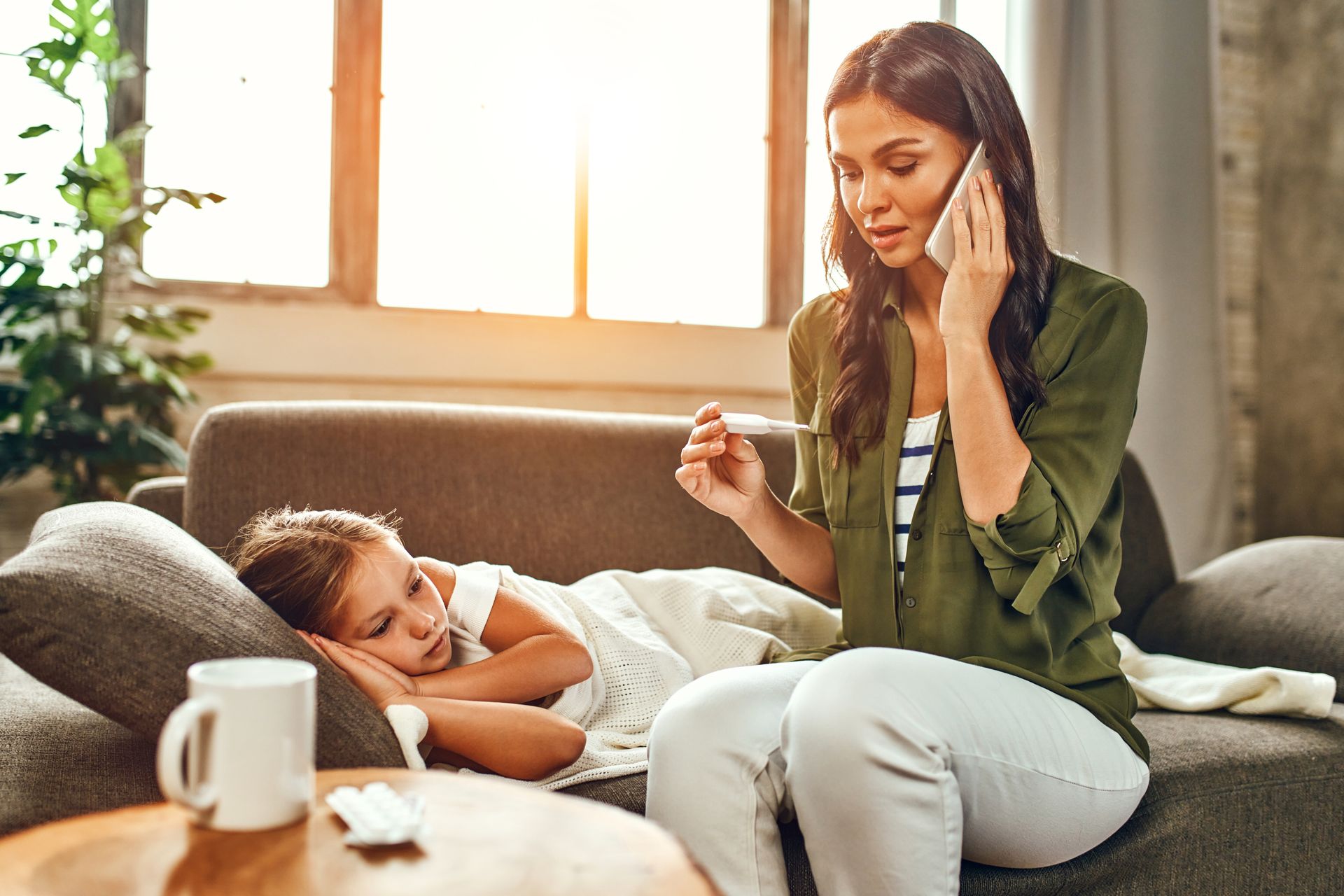 A woman is sitting on a couch talking on a cell phone while a little girl is laying on the couch.