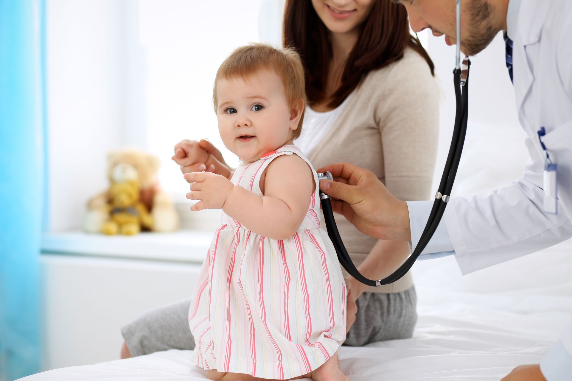 A baby girl is on a medical table, her mom is standing next to her. A doctor is checking the baby's heart with a stethoscope.