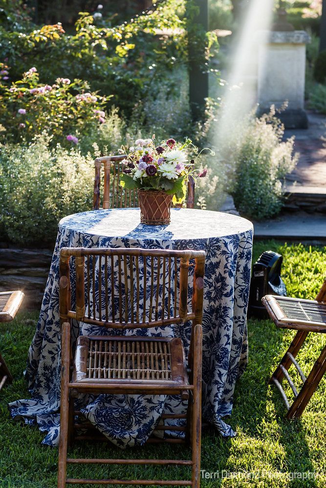 Sunlit garden table with floral tablecloth, chair, and potted flowers amid greenery