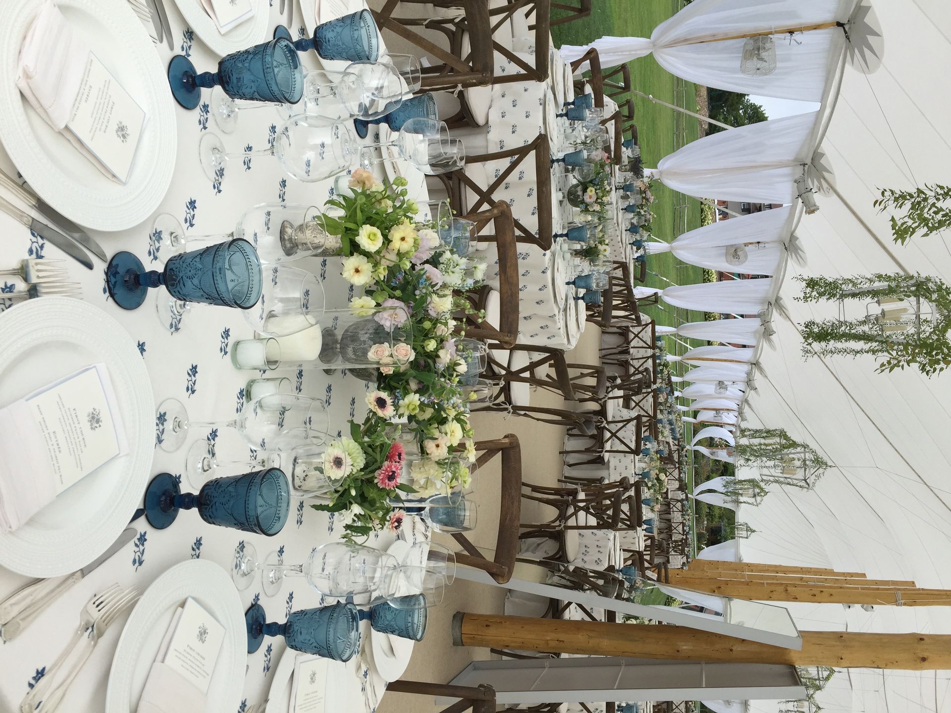 Outdoor banquet setup with white-draped tables, blue glassware, floral centerpieces, and string lights under a canopy