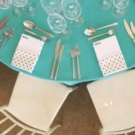 Top-down view of a turquoise table set with glassware, silverware, and polka-dot napkins beside white chairs