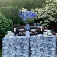 Outdoor buffet table with blue floral tablecloths, white dishes, and purple and white floral arrangements