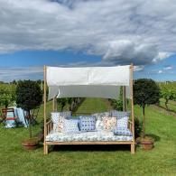 White canopy daybed on a grassy lawn with pillows and an orchard under a cloudy sky.