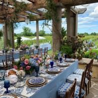 Outdoor dining table under a pergola with blue and floral table settings overlooking a garden and water view