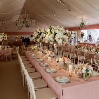 Elegant wedding reception table under a white tent with blush tablecloth, floral centerpiece, and chandeliers