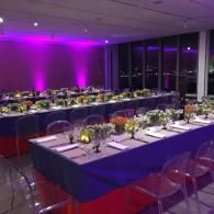 Purple-lit banquet table setup in a modern room with long tables, glassware, and floral centerpieces
