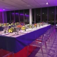 Purple-lit banquet table with white tablecloth, floral centerpieces, and clear chairs by floor-to-ceiling windows