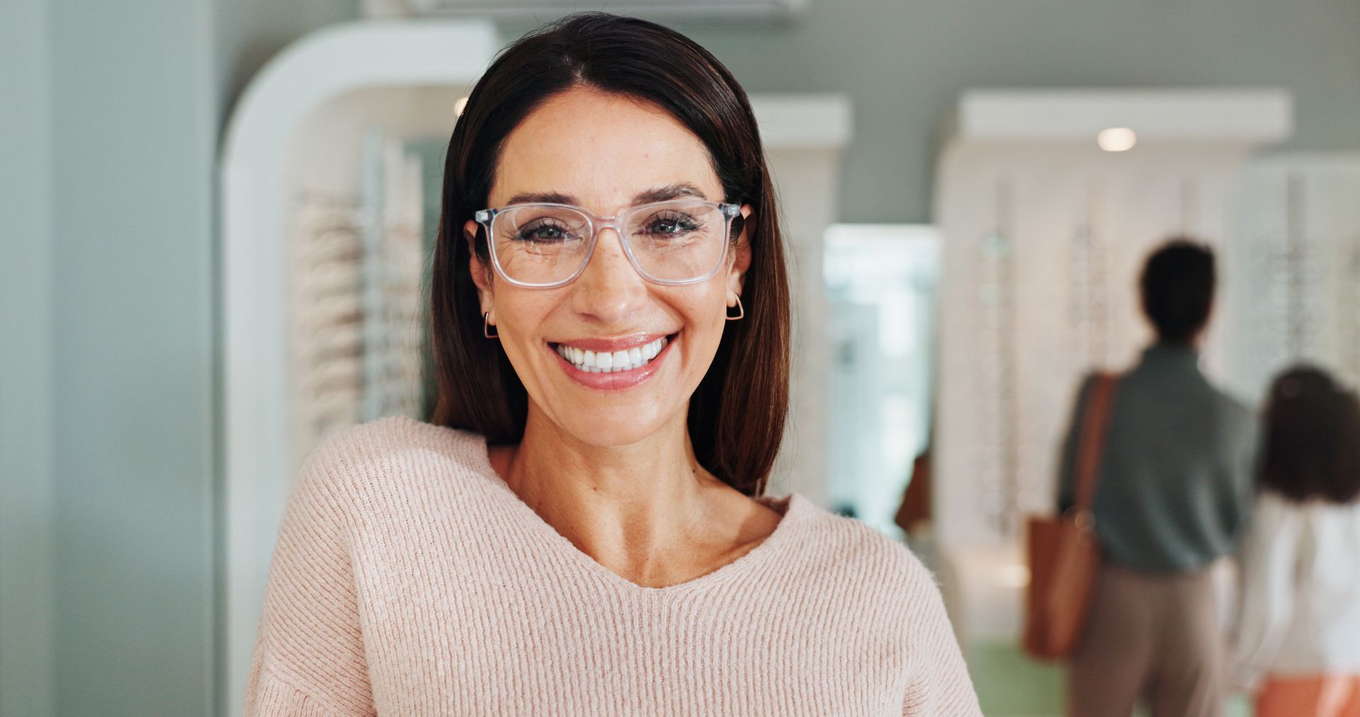 Smiling woman in glasses and pink sweater standing in a bright hallway with blurred figures behind her Smiling woman in glasses and pink sweater standing in a bright hallway with blurred figures behind her