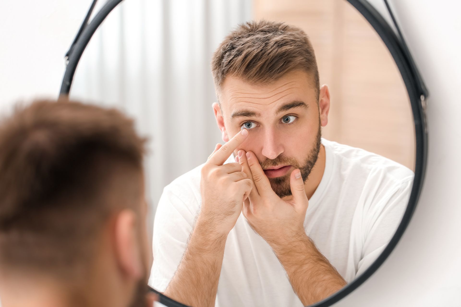 Man in white shirt checking skin in a round mirror, touching his face close up