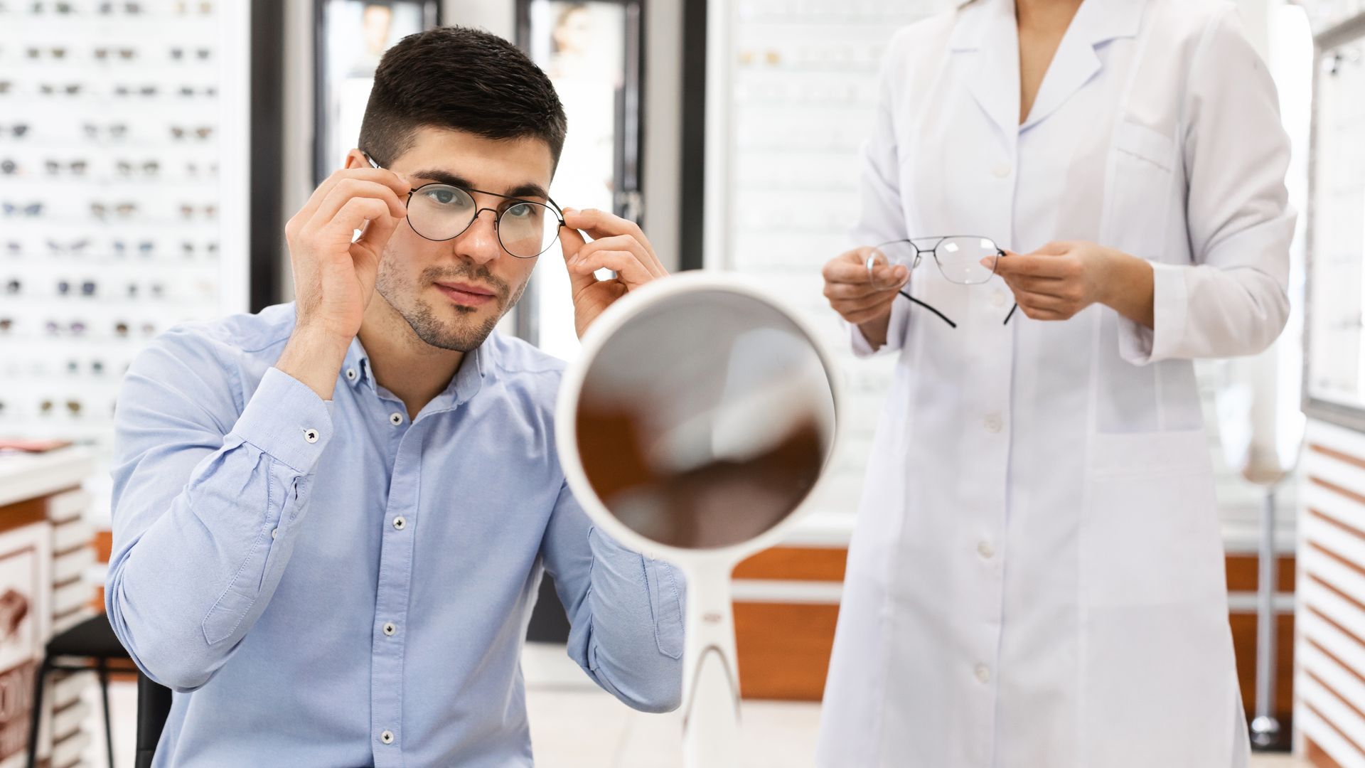 Man adjusting glasses in an eye exam room while an optometrist holds frames nearby Man adjusting glasses in an eye exam room while an optometrist holds frames nearby