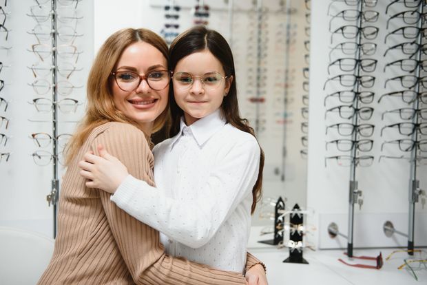 Two women smiling in an eyeglasses store, standing in front of rows of frames.