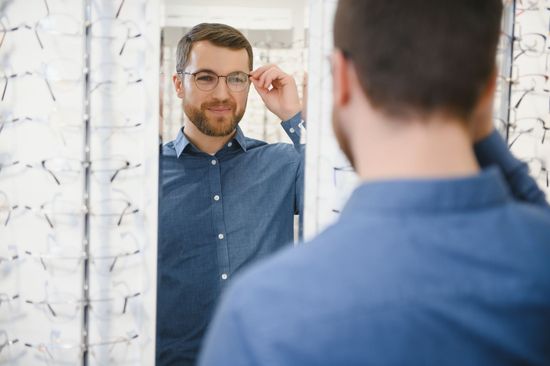 Man adjusting glasses while looking in a mirror, with his back to the camera in a bright room.