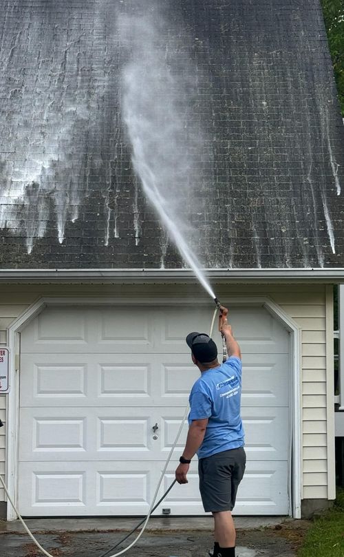 Person pressure-washes a house roof and siding with a long spray wand.