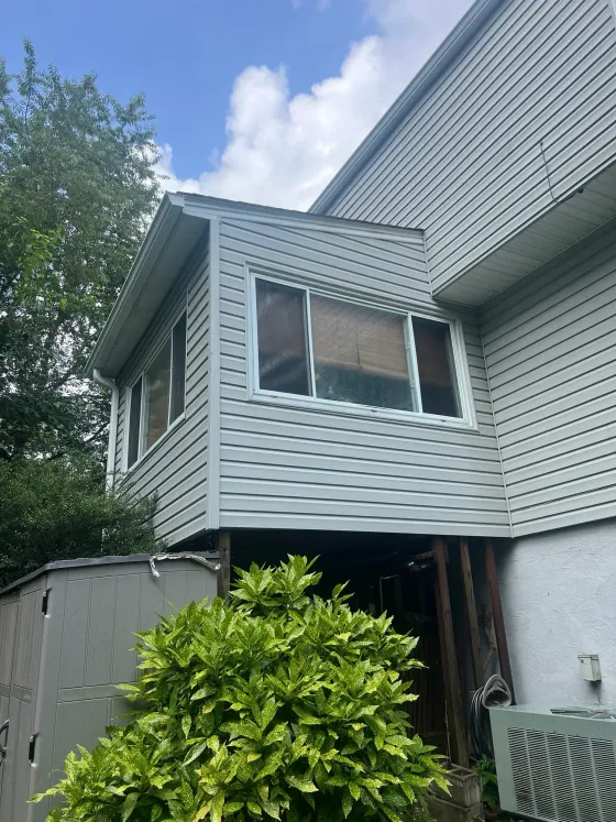 Suburban house exterior with gray siding, a large window, and shrubs beneath a cloudy sky